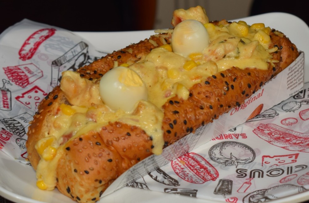 Perro Granjero (Pan brioche, salchicha, Tocineta Ahumada, Pollo en salsa de quesos, Maíz tierno, Queso, Papitas, Cebolla picada)